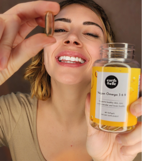 a woman wearing a beige top holding a softel of vegan omega supplement in one hand and the bottle in the other, smiling while doing so.