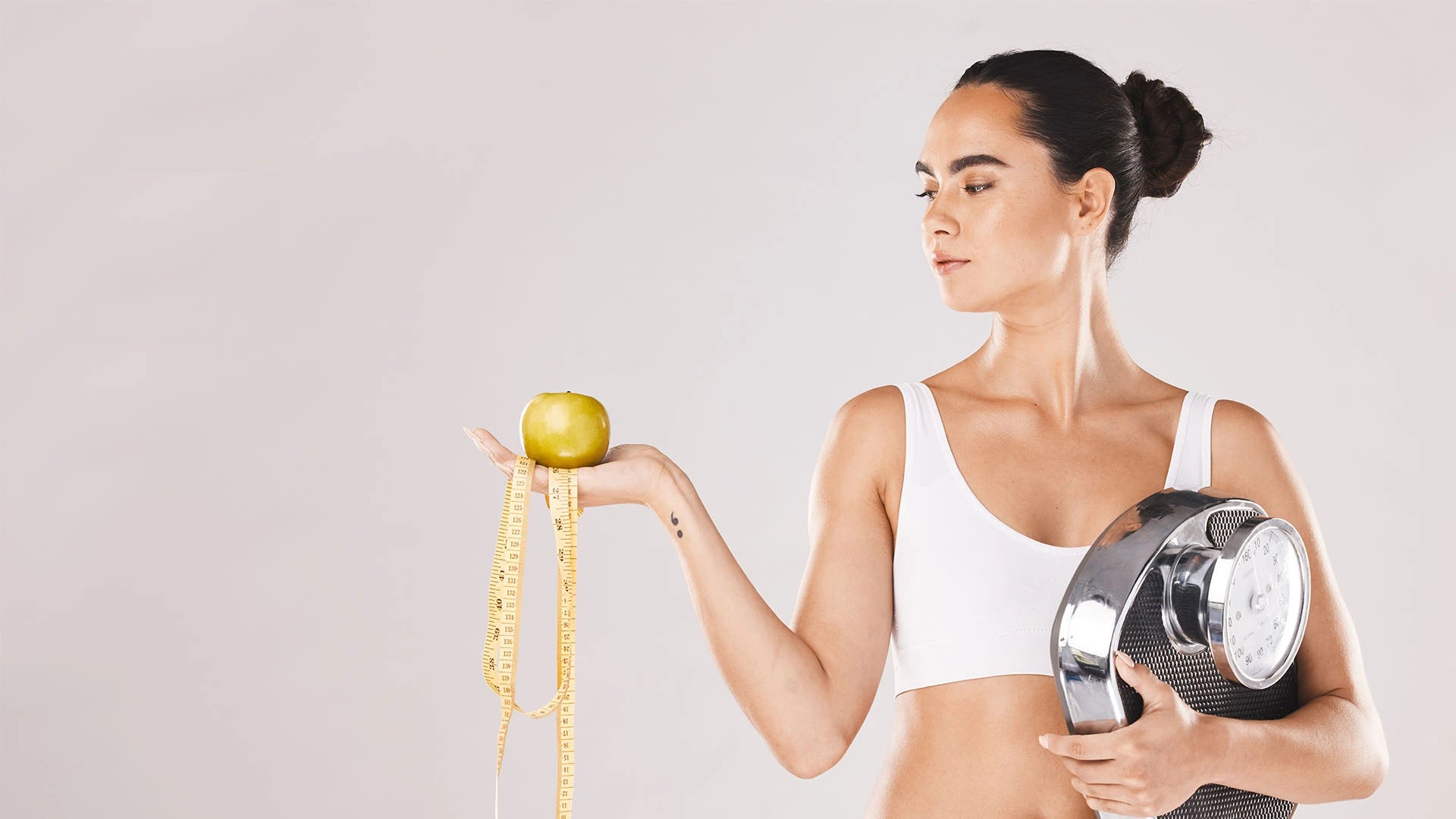 Fit woman in a white sports bra holding a green apple with a yellow measuring tape draped over her hand, while clutching a silver analog scale against her side. Symbolizes healthy weight loss, balanced nutrition, and body confidence through mindful eating and fitness