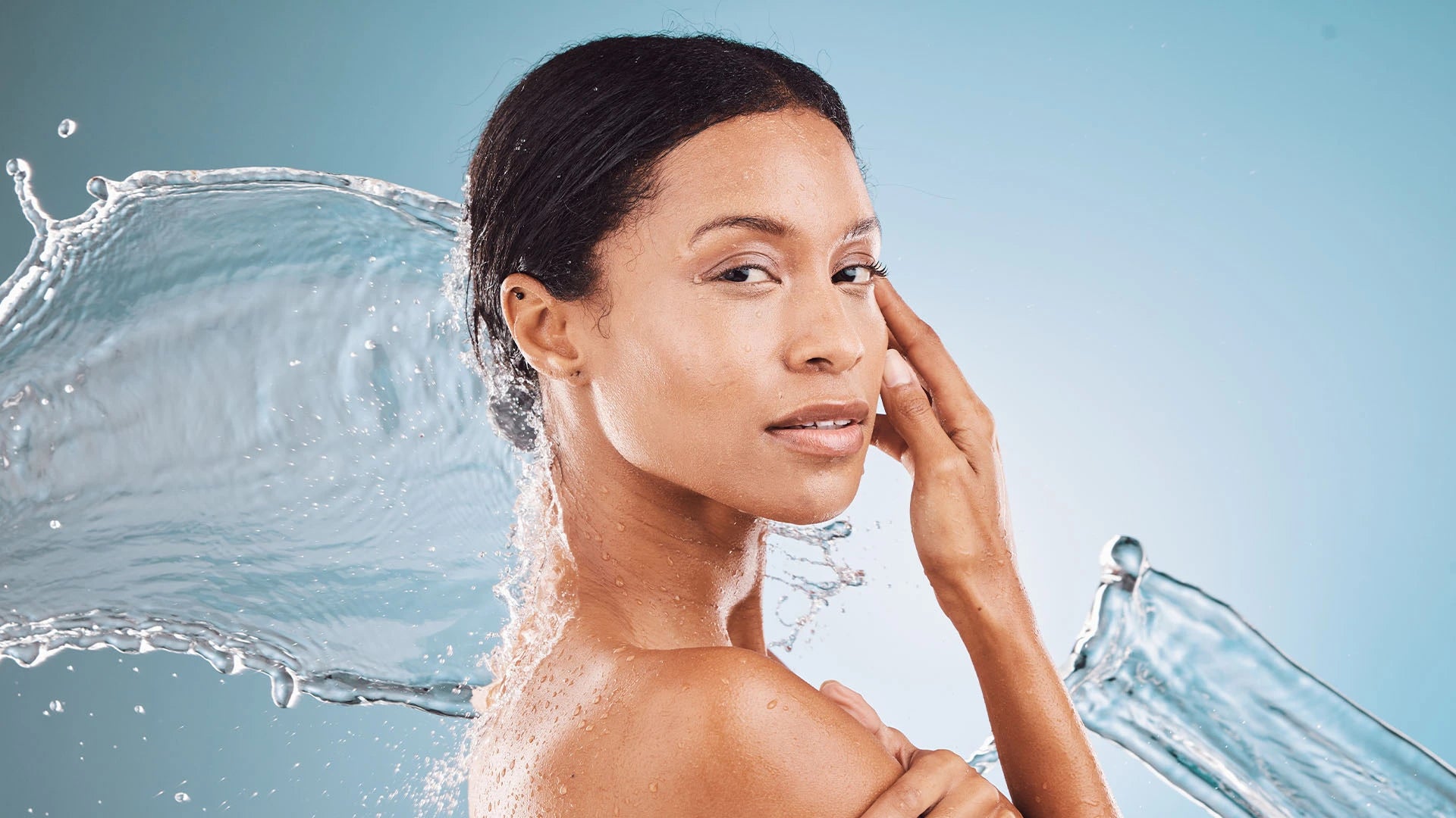Confident woman with dewy, radiant skin standing against a light blue background as a splash of water flows behind her, symbolizing intense hydration and skin moisture balance. Represents a skincare routine focused on hydration, glow, and natural beauty